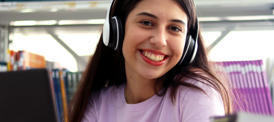 Imagem mostra uma jovem estudante branca sorrindo para a foto. Ela usa fone de ouvido, um notebook, uma camiseta lilás e está em um ambiente que parece ser uma biblioteca (Ciência de dados)