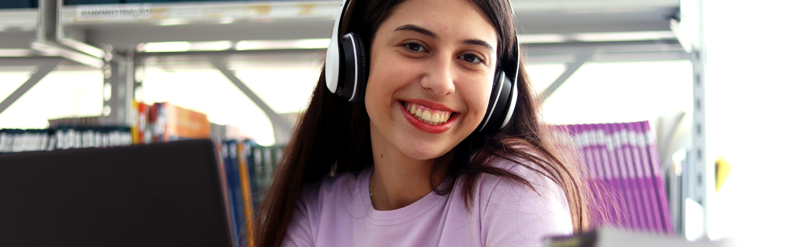 Imagem mostra uma jovem estudante branca sorrindo para a foto. Ela usa fone de ouvido, um notebook, uma camiseta lilás e está em um ambiente que parece ser uma biblioteca (Ciência de dados)