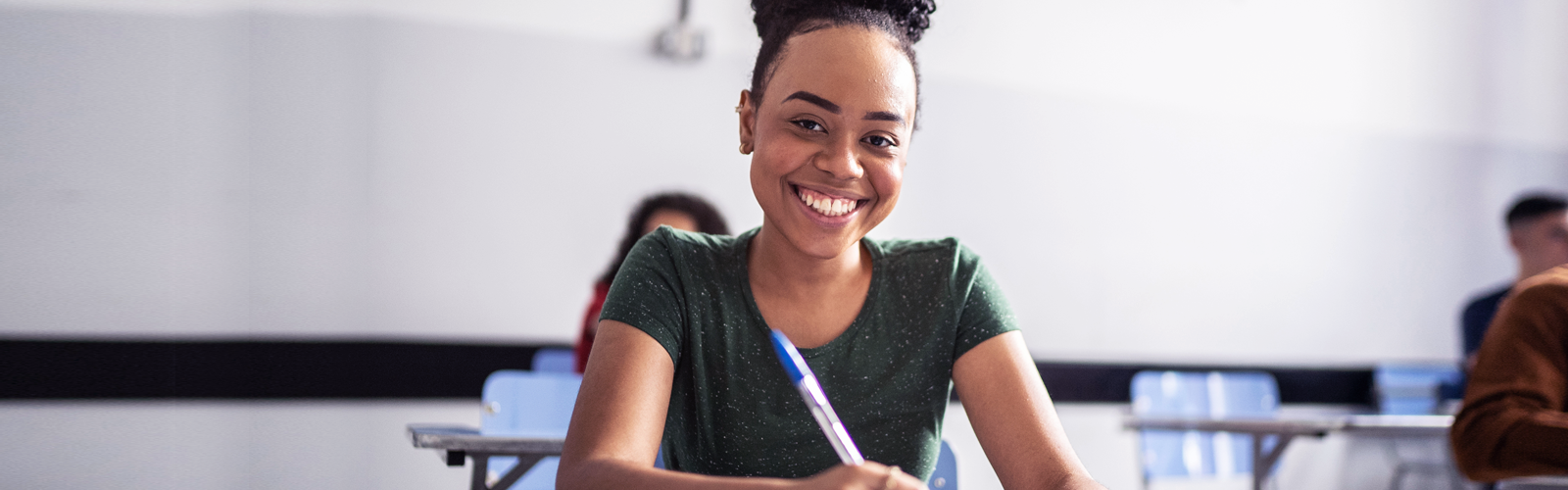 Imagem mostra uma jovem estudante negra sentada em uma cadeira em sala de aula. Ela sorri para a foto e segura uma caneta na mão direita (Novo Ensino Médio)