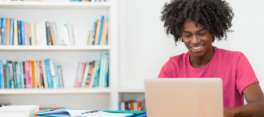 Imagem mostra um jovem negro utilizando um notebook. Ele usa camiseta rosa, está em um ambiente que parece ser uma sala de estudos. O notebook está sobre uma mesa branca. Ao fundo, é possível ver uma estante com livros