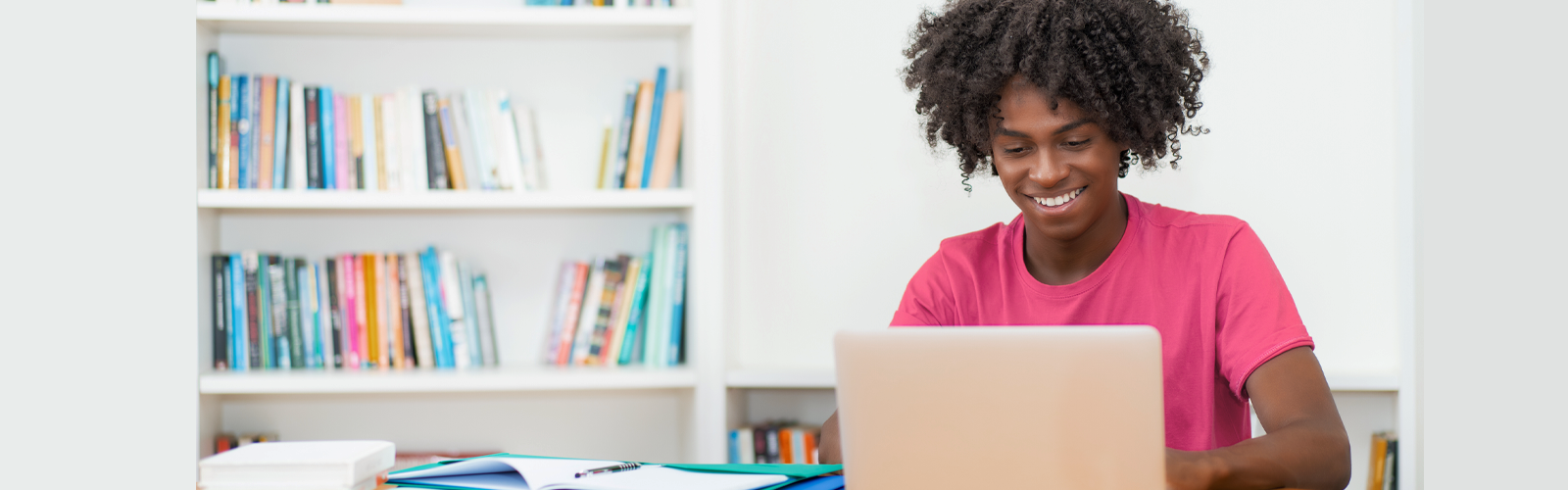 Imagem mostra um jovem negro utilizando um notebook. Ele usa camiseta rosa, está em um ambiente que parece ser uma sala de estudos. O notebook está sobre uma mesa branca. Ao fundo, é possível ver uma estante com livros