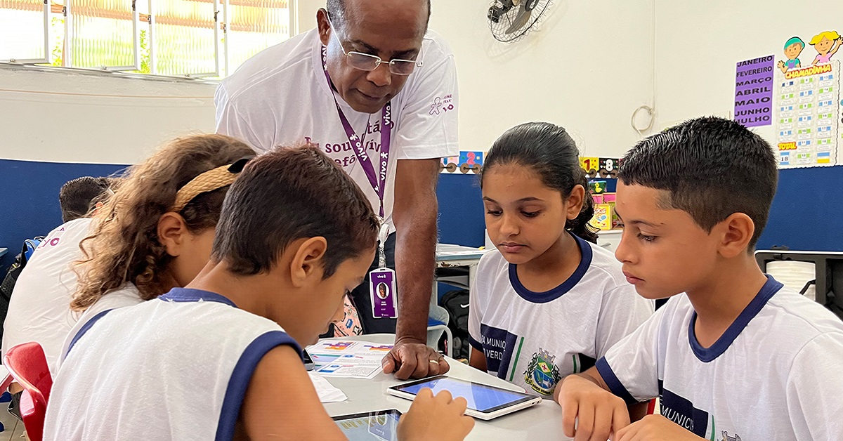 Imagem mostra um voluntário negro orientando quatro alunos em uma sala de aula. O voluntário usa óculos. Os alunos estão sentados e observam tablets que estão em cima da mesa.