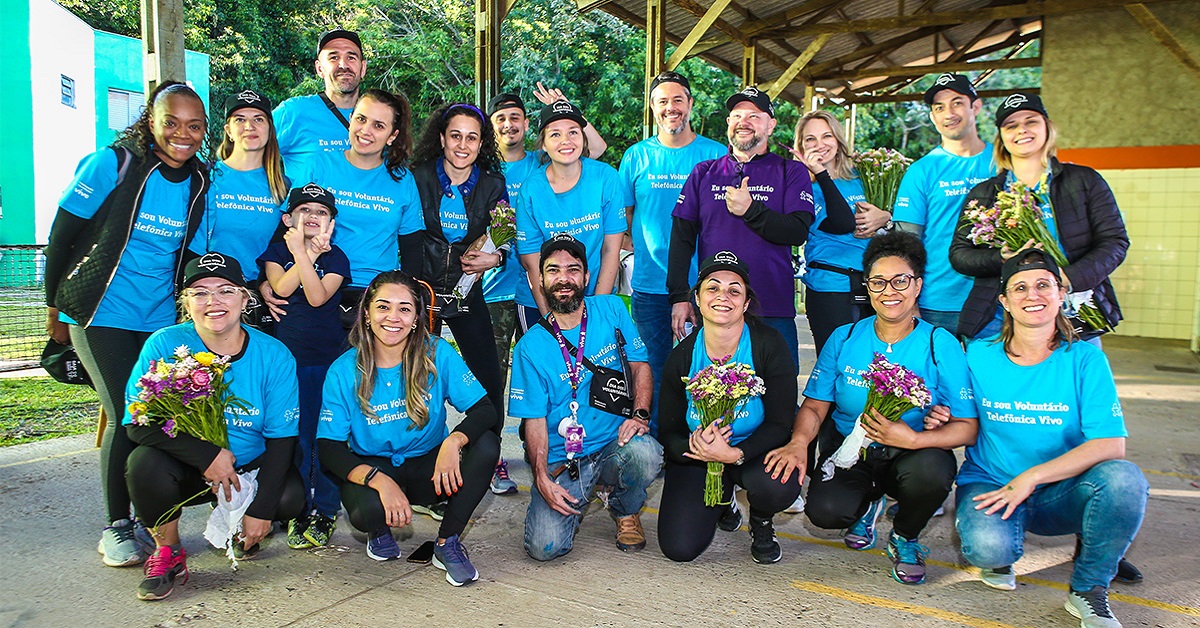 Imagem mostra grupo de 19 voluntários, entre homens e mulheres, reunidos em uma instituição. Eles usam camiseta azul com a frase Eu sou voluntário Telefônica