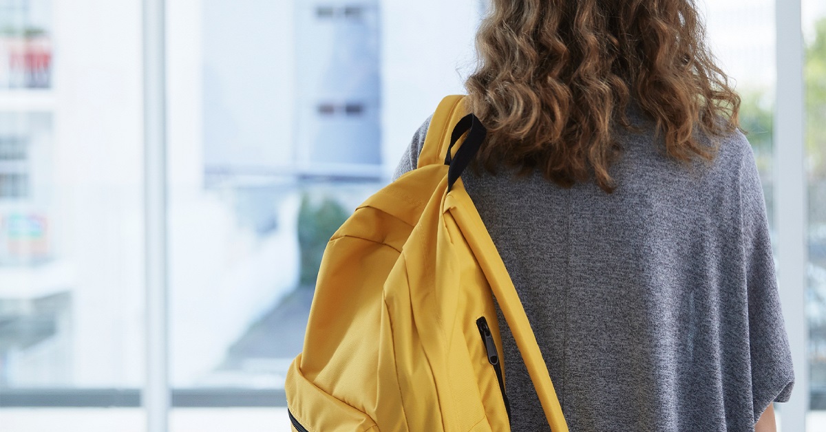 Aluna chegando na escola, de costas, de mochila amarela.