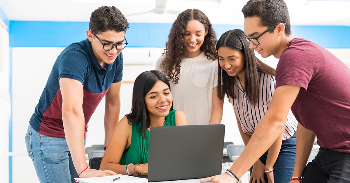 Imagem mostra uma professora usando um notebook, enquanto quatro jovens estudantes estão ao seu lado acompanhando o que ela está fazendo. Todos são brancos e estão sorrindo