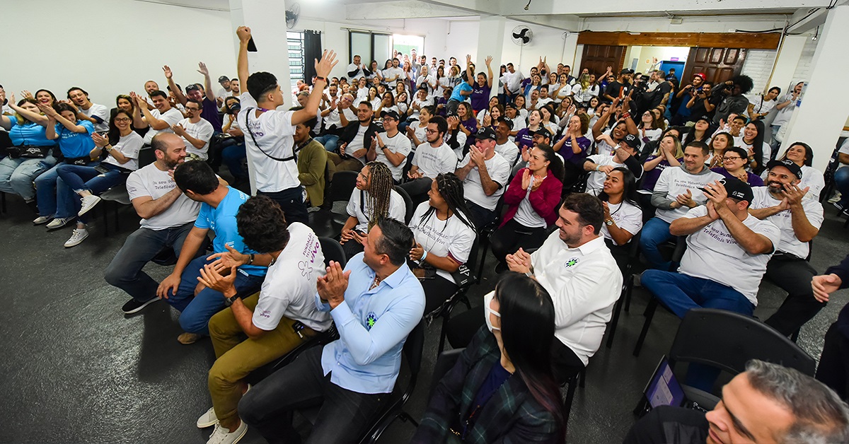 Imagem mostra grupo de voluntários e de estudantes reunidos em um espaço fechado durante o Dia dos Voluntários. As pessoas estão aplaudindo a fala de um jovem que está em pé à frente de todos