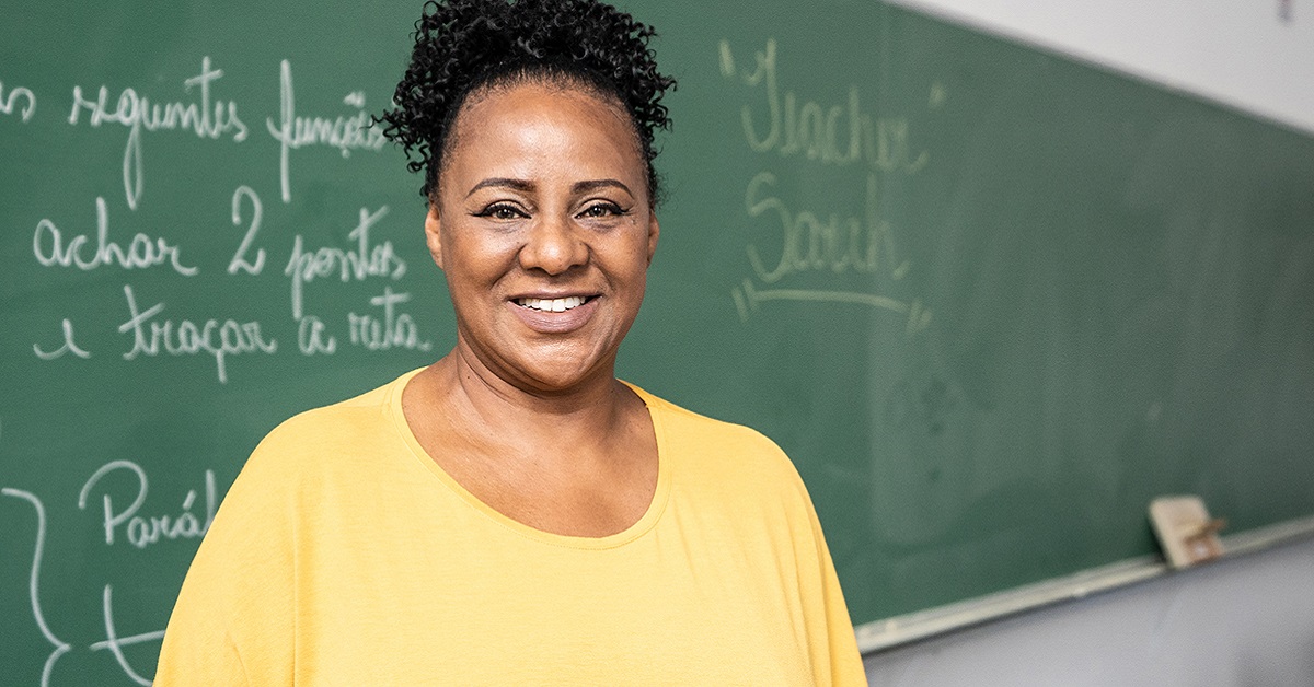 Imagem mostra uma professora negra em sala de aula, sorrindo para a foto. Ela usa blusa amarela e cabelos presos. Ao fundo, há uma lousa