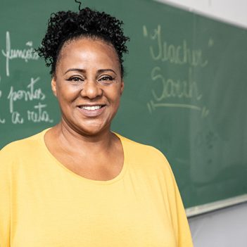 Imagem mostra uma professora negra em sala de aula, sorrindo para a foto. Ela usa blusa amarela e cabelos presos. Ao fundo, há uma lousa