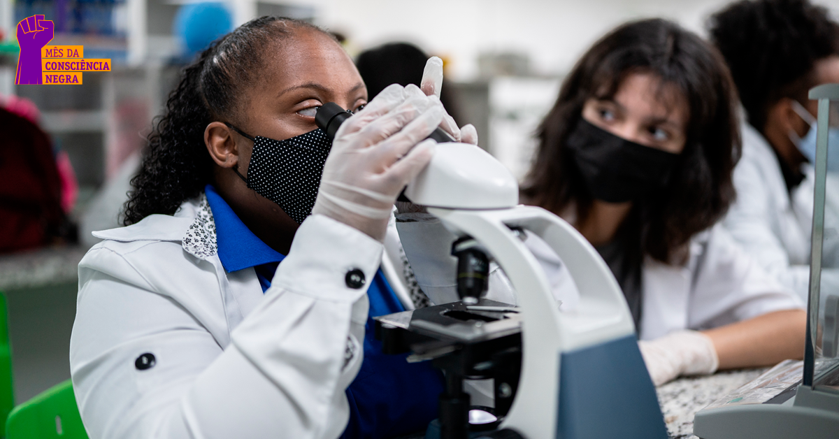 Cientista negra, vestida com jaleco branco, olha em um visor de mi-croscópio.