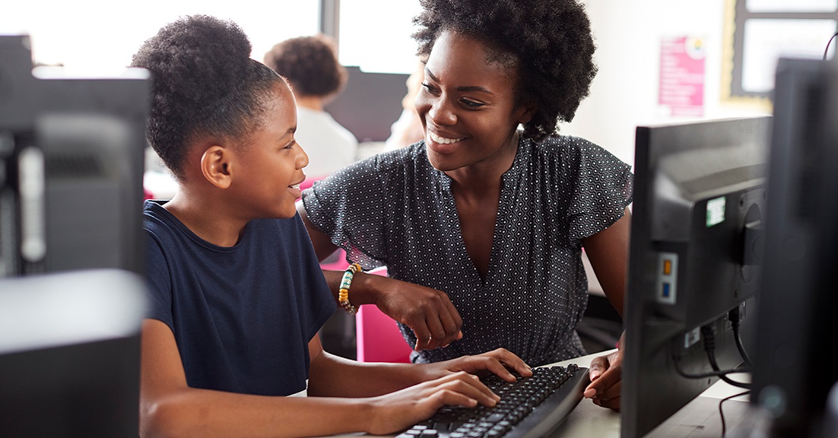 Imagem mostra uma professora negra ao lado de uma aluna negra. Elas estão utilizando um computador