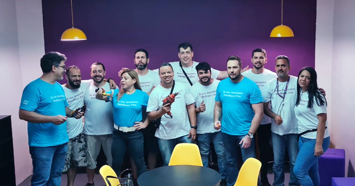 Imagem mostra um grupo de 12 voluntários; são 10 homens e 2 mulheres. Eles estão reunidos em uma sala, ao fundo há uma parede na cor roxa. Eles usam camisetas com a frase sou voluntário telefônica