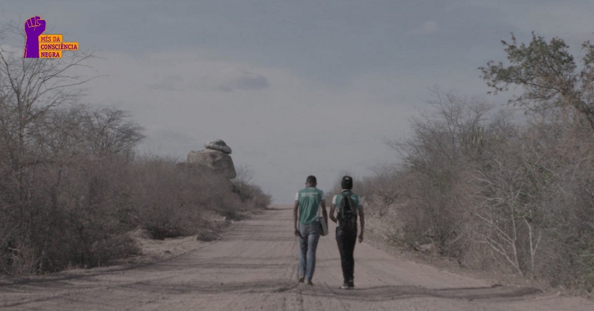 A imagem mostra dois jovens de costas, andando por uma estrada de terra, numa paisagem do semiárido do Ceará. O rapaz da esquerda veste calça jeans e camiseta do uniforme escolar verde e está com um caderno na mão direita. O rapaz da direita veste calça jeans e camiseta verde do uniforme escolar e tem uma mochila preta nas costas