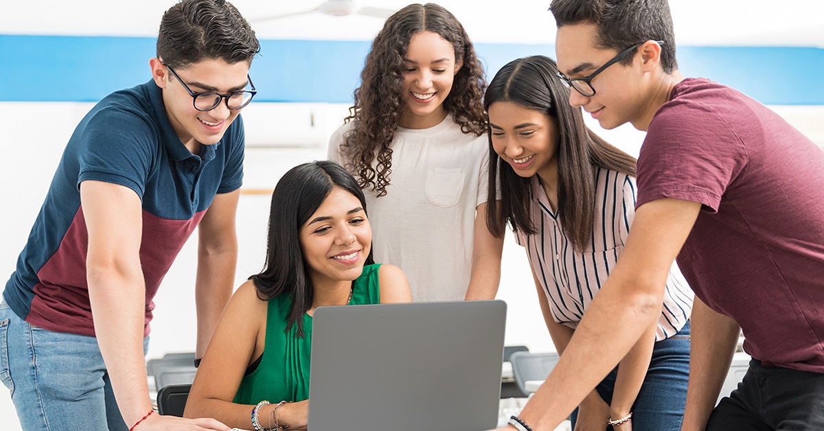 Imagem mostra um grupo de jovens estudantes ao redor de uma professora que está sentada usando um computador. Todos estão em uma sala de aula