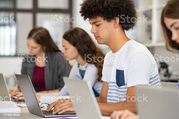 College student using laptop in class during computer lesson. Side view of guy sitting in a row studying in classroom on pc. Black high school student studying on laptop in university library.