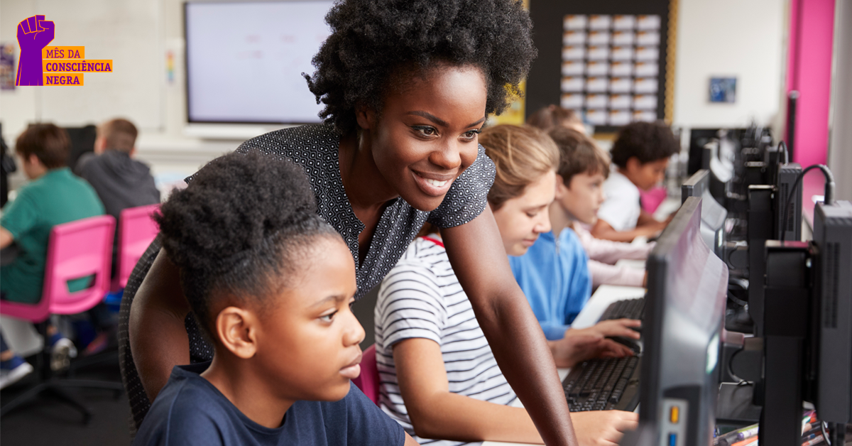 Imagem mostra uma sala de computadores com adolescentes. Em primeiro plano está uma estudante olhando para a tela do computador. Atrás dela, em pé está a professora, sorrindo e mexendo no mouse.