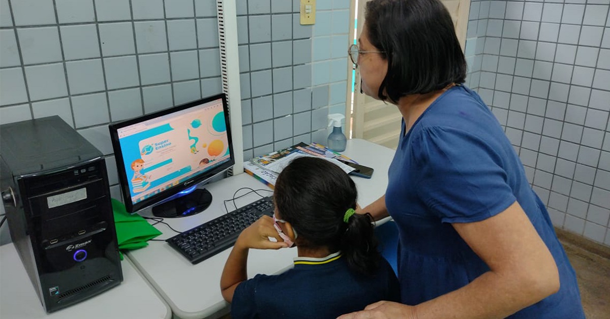 Imagem mostra uma professora auxiliando uma aluna a usar um computador em sala de aula