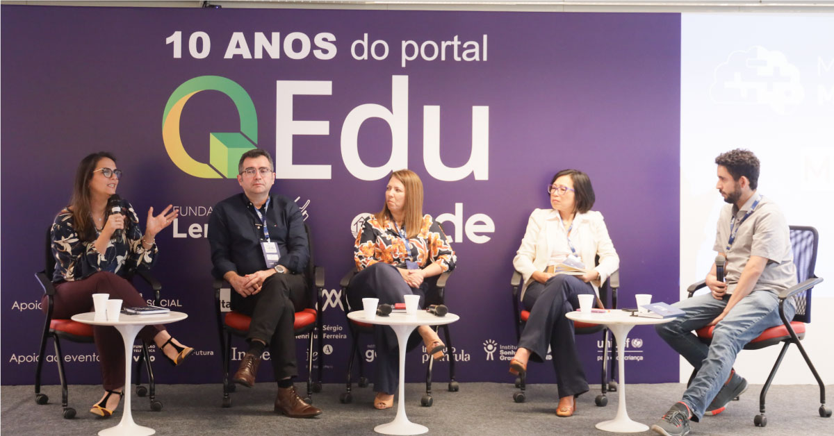 Imagem mostra o palco do evento, onde estão sentados em cadeiras, lado a lado, três palestrantes mulheres e dois homens. Há três mesas de centro no palco e ao fundo um painel com a frase 10 anos do portal QEdu