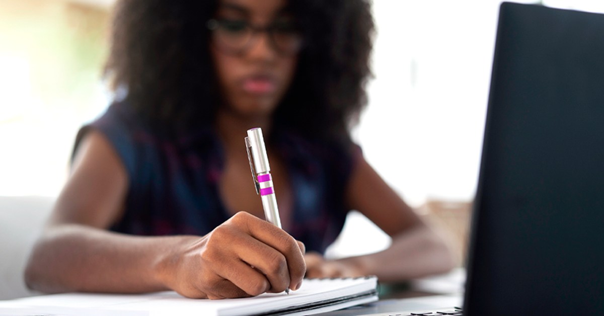 Imagem mostra uma educadora negra escrevendo em um caderno. À sua frente, há um notebook preto