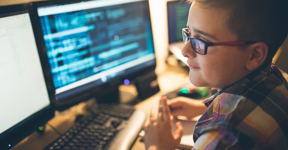 Adolescente usa óculos e camisa xadrez. Está em sentado em frente ao computador, com duas telas.