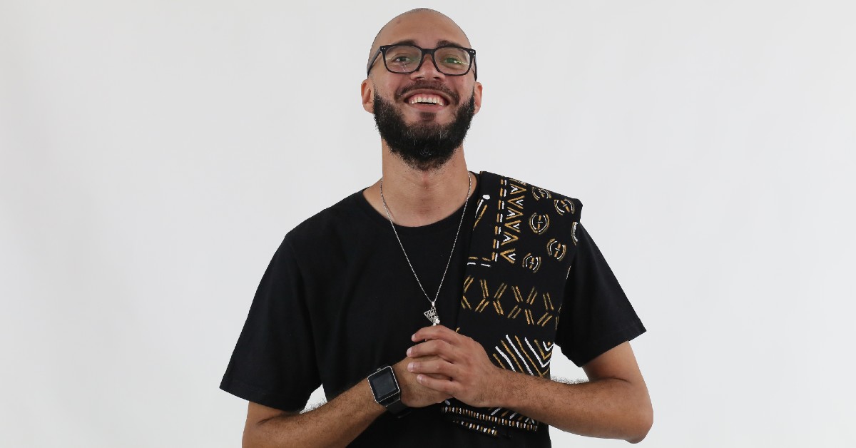 A foto mostra o escritor Israel Neto, fundador da Editora Kitembo. Ele veste camiseta preta, colar e lenço com símbolos e estampas africanas.