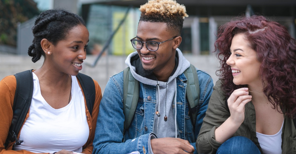 A imagem mostra três jovens de ensino médio. A jovem da direita tem cabelo vermelho, veste blusa de manga comprida verde, camiseta branca e calça jeans e traz a mão levantada na altura do queijo. O jovem do meio usa óculos e tem cabelos pintados de loiro. Está vestindo blusa de moletom cinza, jaqueta jeans e mochila cinza nas costas. A jovem da ponta esquerda tem cabelo preto e com trança, veste camiseta branca, blusa de frio laranja, mochila preta nas costas e está com alguns papeis na mão direita. Os três estão sorrindo.