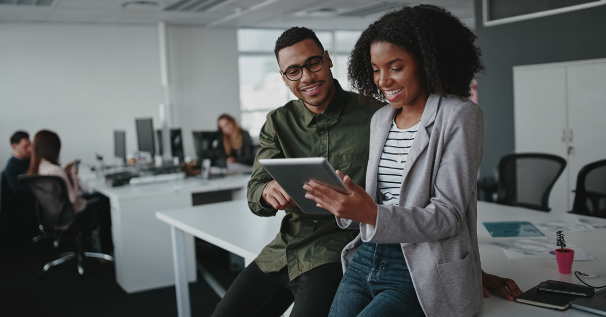 A imagem mostra dois jovens profissionais, um homem e uma mulher, no escritório, conversando enquanto utilizam um laptop, nas mãos da jovem mulher