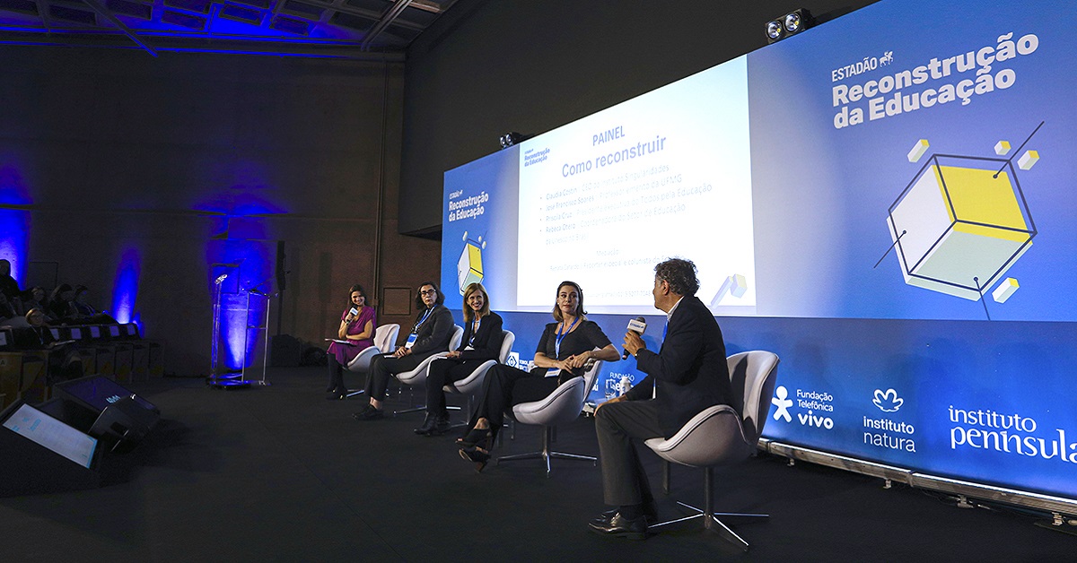 Imagem mostra o palco do evento, com cinco palestrantes sentados em cadeiras sobre o palco