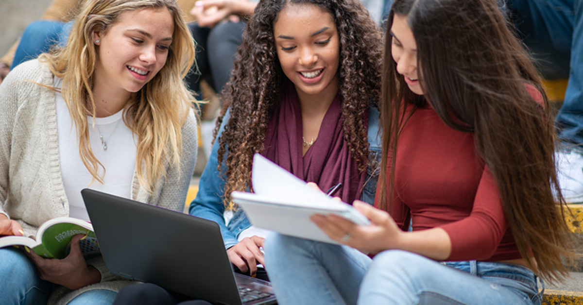 Imagem mostra três estudantes sentadas lado a lado. Uma delas segura um notebook e outra está com um papel na mão, que está sendo observado por todas