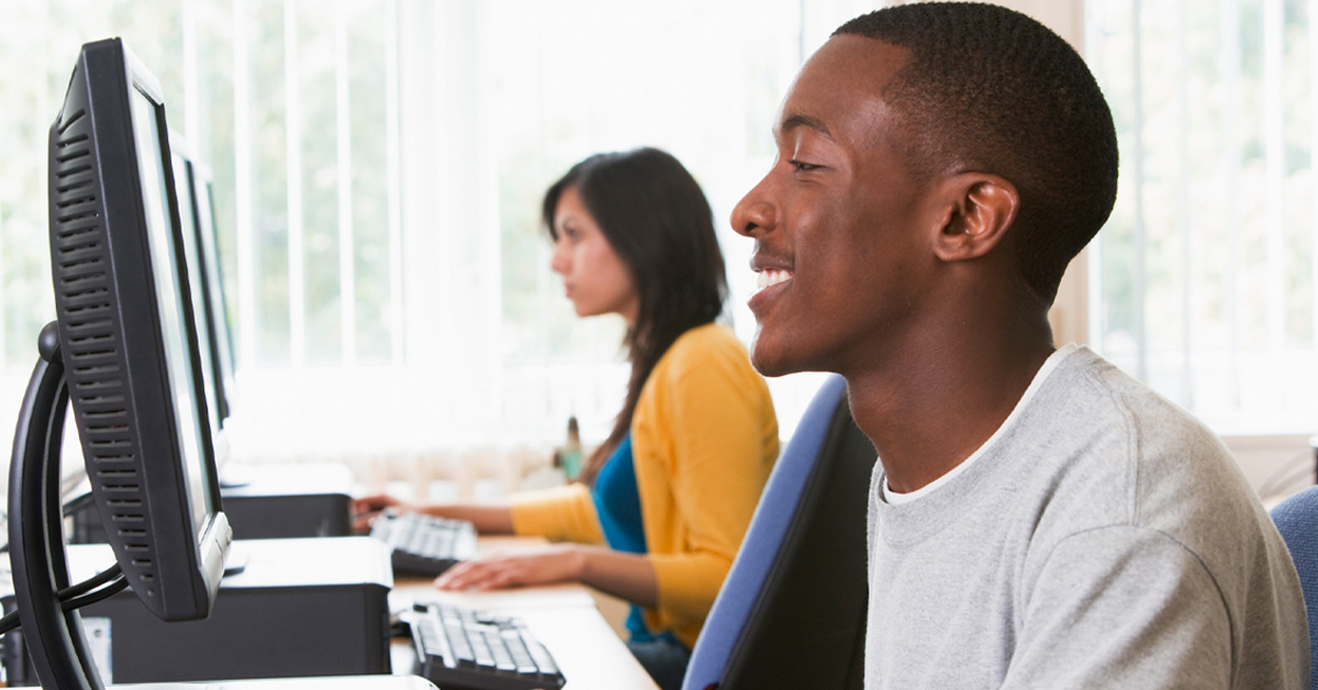 Imagem mostra um jovem estudante negro sentado de frente a um computador. Ao fundo, é possível ver uma estudante, branca, também em um computador