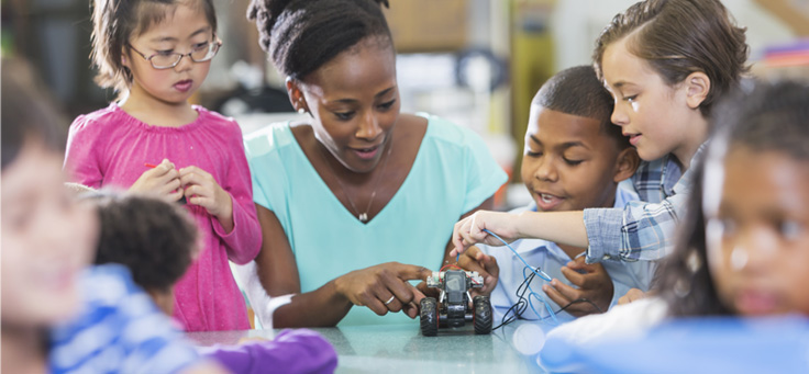 Imagem mostra uma professora negra com alunos brancos, negros e orientais. Eles estão manuseando um objeto de robótica