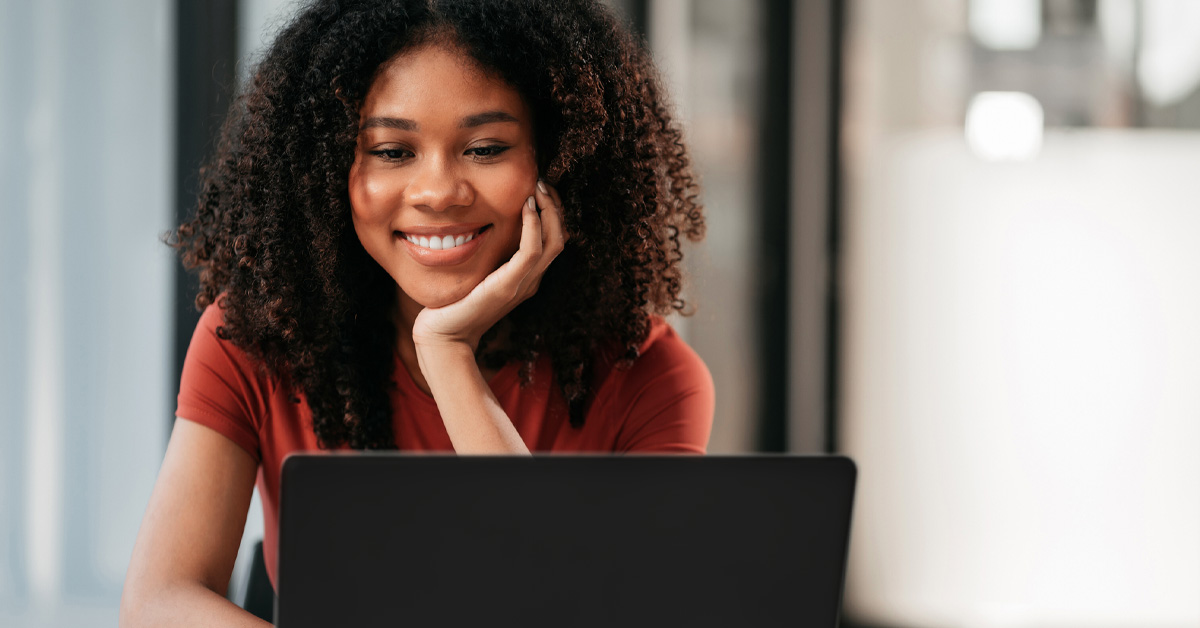 Imagem mostra uma jovem negra, ela tem cabelos compridos, solto, e ela está olhando para um notebook, com a mão esquerda no queixo