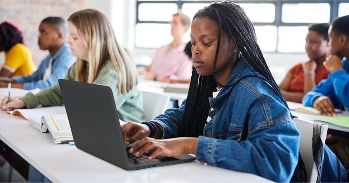 A imagem mostra uma estudante negra digitando em seu notebook. Ela está vestindo jaqueta jeans. Ao seu lado, estão outros estudantes em uma bancada de estudos.