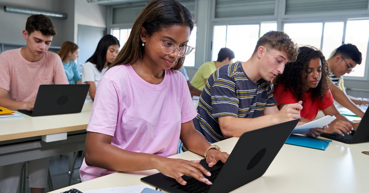 A imagem mostra um grupo diverso de estudantes adolescentes. Eles estão sentados em duas bancadas de estudos com cadernos, canetas e notebooks. Em primeiro plano, a imagem mostra quatro estudantes: duas meninas e dois meninos. Em segundo plano, é possível ver mais quatro estudantes, também dois meninos e duas meninas.