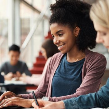 A imagem mostra uma estudante negra que veste camiseta azul e blusa vermelha sorrindo e digitando em um notebook preto. Em desfoca, uma estudante loira que veste camisa jeans está com um lápis na mão.