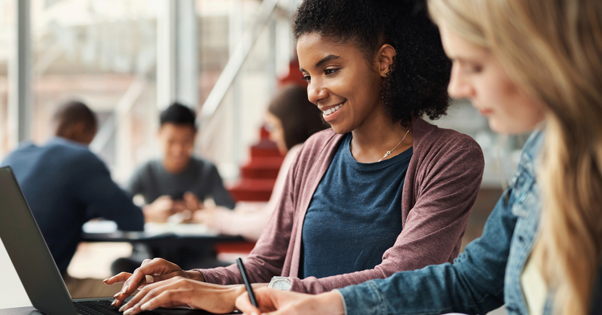 A imagem mostra uma estudante negra que veste camiseta azul e blusa vermelha sorrindo e digitando em um notebook preto. Em desfoca, uma estudante loira que veste camisa jeans está com um lápis na mão.