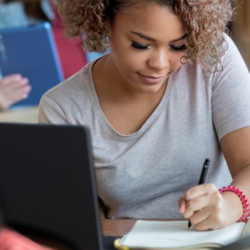 Imagem mostra uma estudante negra, com cabelo ondulado, solto, em uma sala de aula, sentada em uma cadeira, escrevendo em um caderno com a mão esquerda