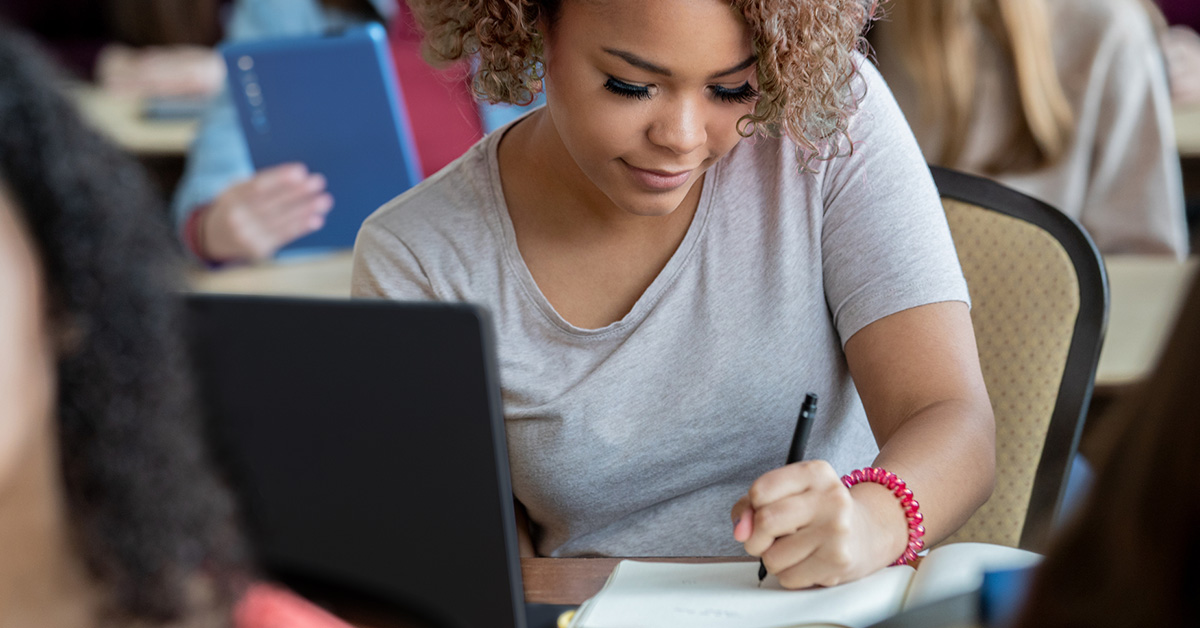 Imagem mostra uma estudante negra, com cabelo ondulado, solto, em uma sala de aula, sentada em uma cadeira, escrevendo em um caderno com a mão esquerda