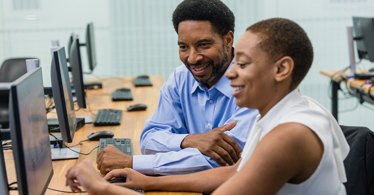 Imagem mostra dois educadores negros, sendo um homem e uma mulher, sentados em uma sala em que estão usando um computador