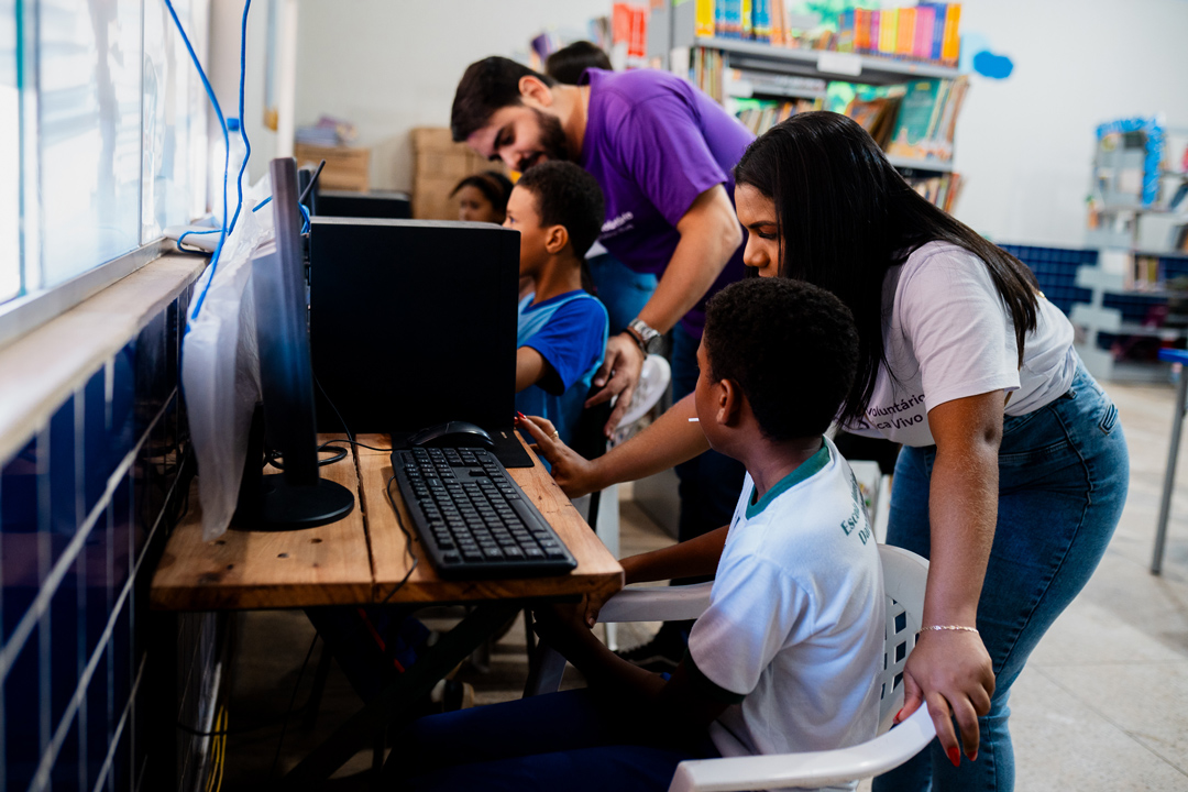Na imagem vemos dois voluntários auxiliando estudantes em uma sala de informática