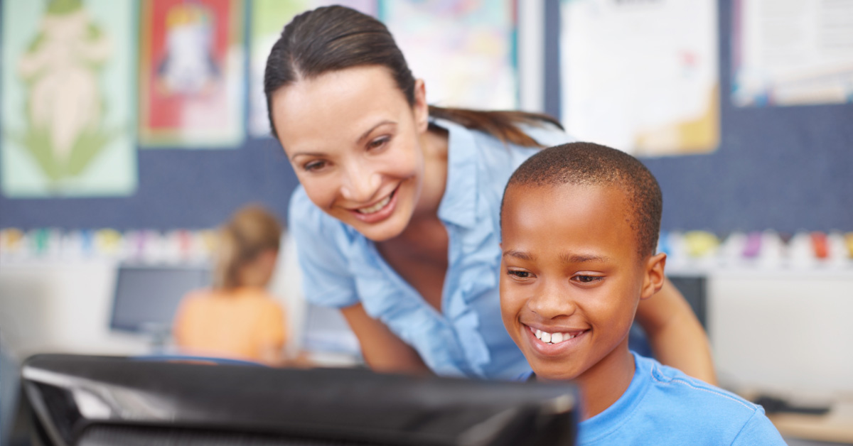 A imagem mostra uma professora adulta de pele clara e cabelos compridos e lisos e um garoto estudante de pele negra e cabelos raspados rentes à cabeça à frente de uma tela de computador. Ao fundo, cartazes cobrem a parede de uma sala da aula.