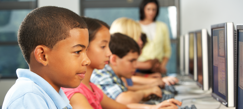 A imagem mostra um grupo de estudantes jovens em frente a computadores. Em primeiro plano, um menino. Ao fundo, a professora que acompanha a aula.