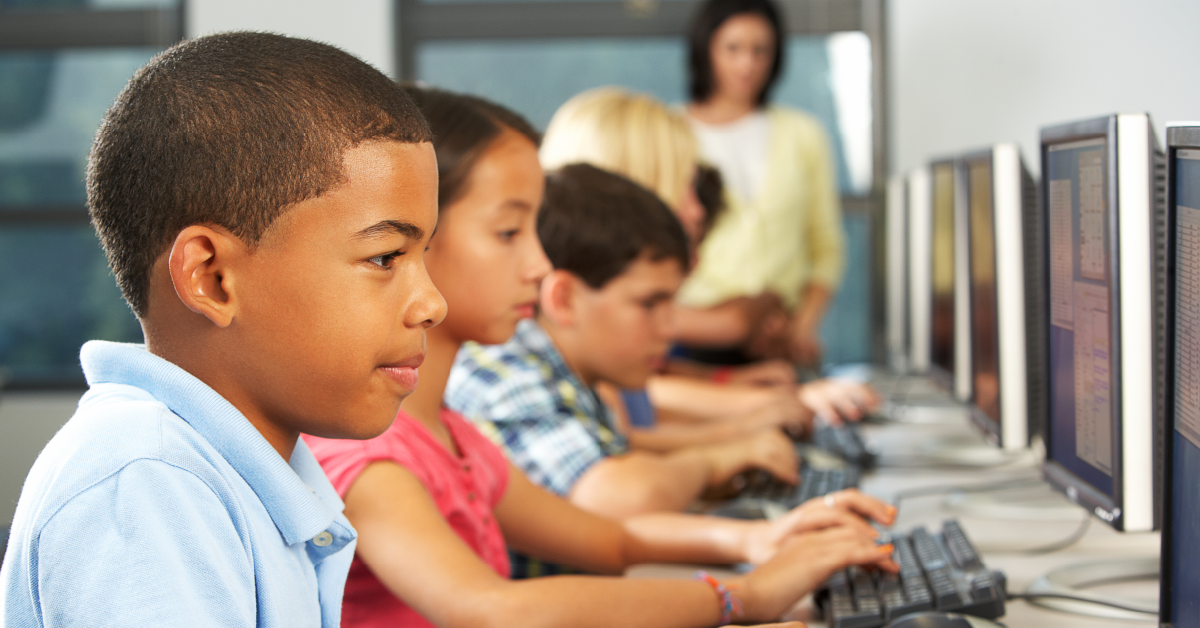 A imagem mostra um grupo de estudantes jovens em frente a computadores. Em primeiro plano, um menino. Ao fundo, a professora que acompanha a aula.