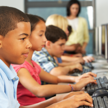 A imagem mostra um grupo de estudantes jovens em frente a computadores. Em primeiro plano, um menino. Ao fundo, a professora que acompanha a aula.