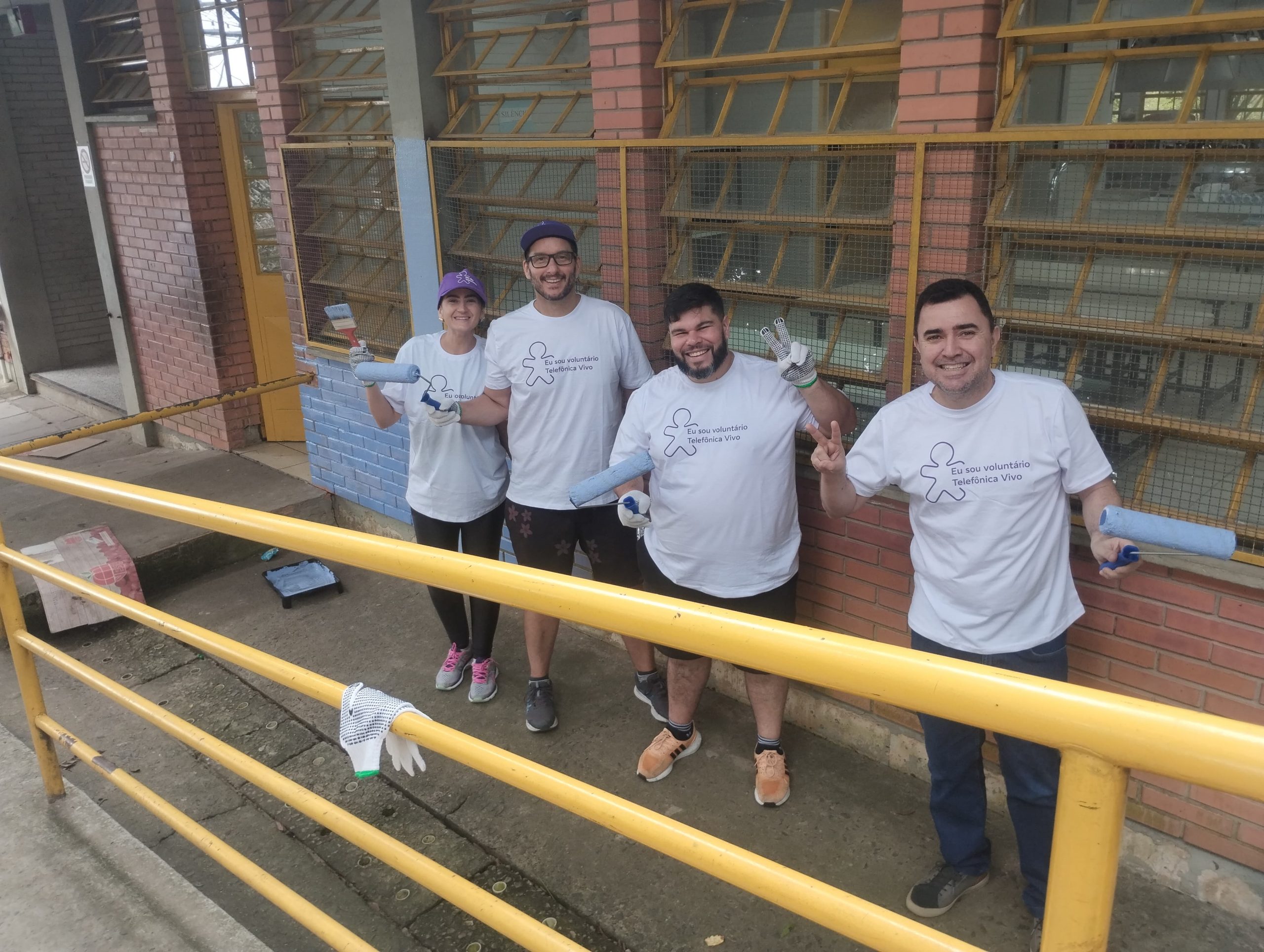 Quatro voluntários, três homens e uma mulher, fazem sinal de vitória e seguram rolos de pintura. Ao fundo, o muro da escola sendo repintado.