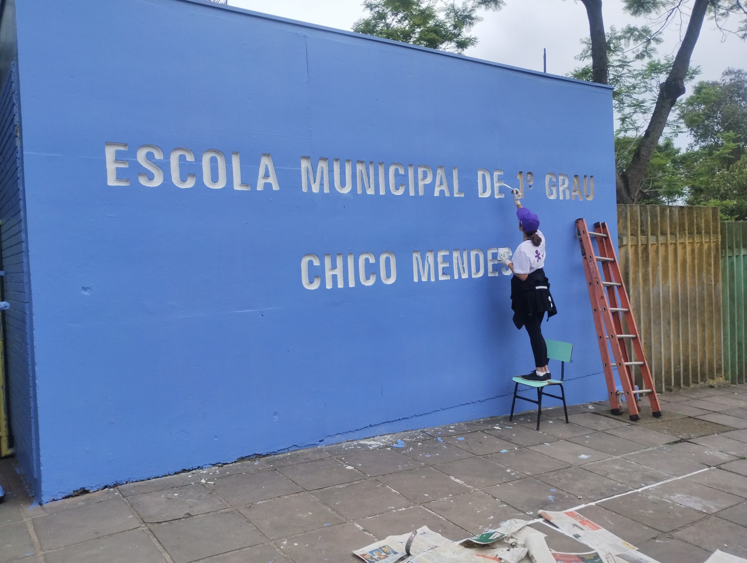 No Dia dos Voluntários RS, mulher faz manutenção na pintura que traz o nome da escola: Escola Municipal de 1º Grau Chico Mendes.