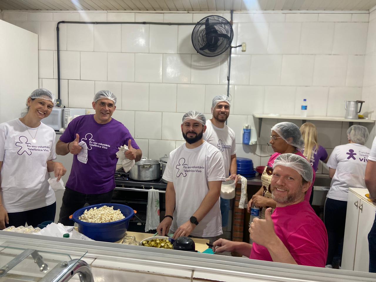 Voluntários posam para foto na cozinha da escola. Diante deles, uma bacia azul cheia de pipoca. Atrás, outro grupo de voluntários cuida da pia.