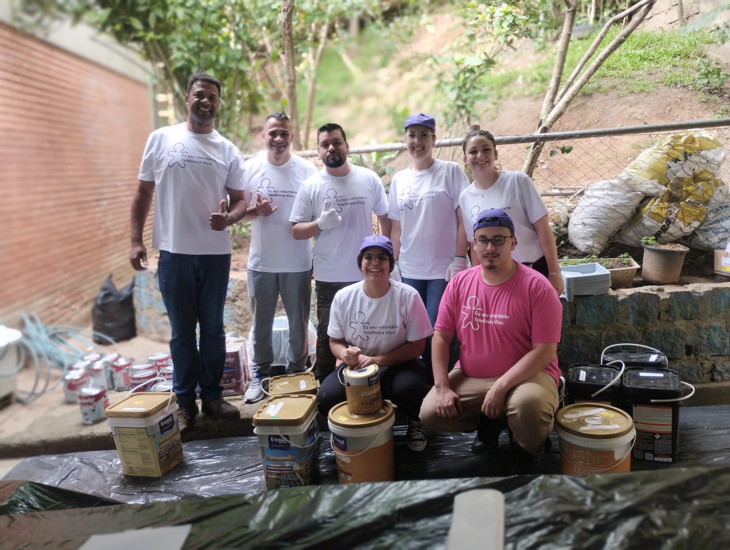 Sete voluntários posam com materiais de pintura no pátio da escola.