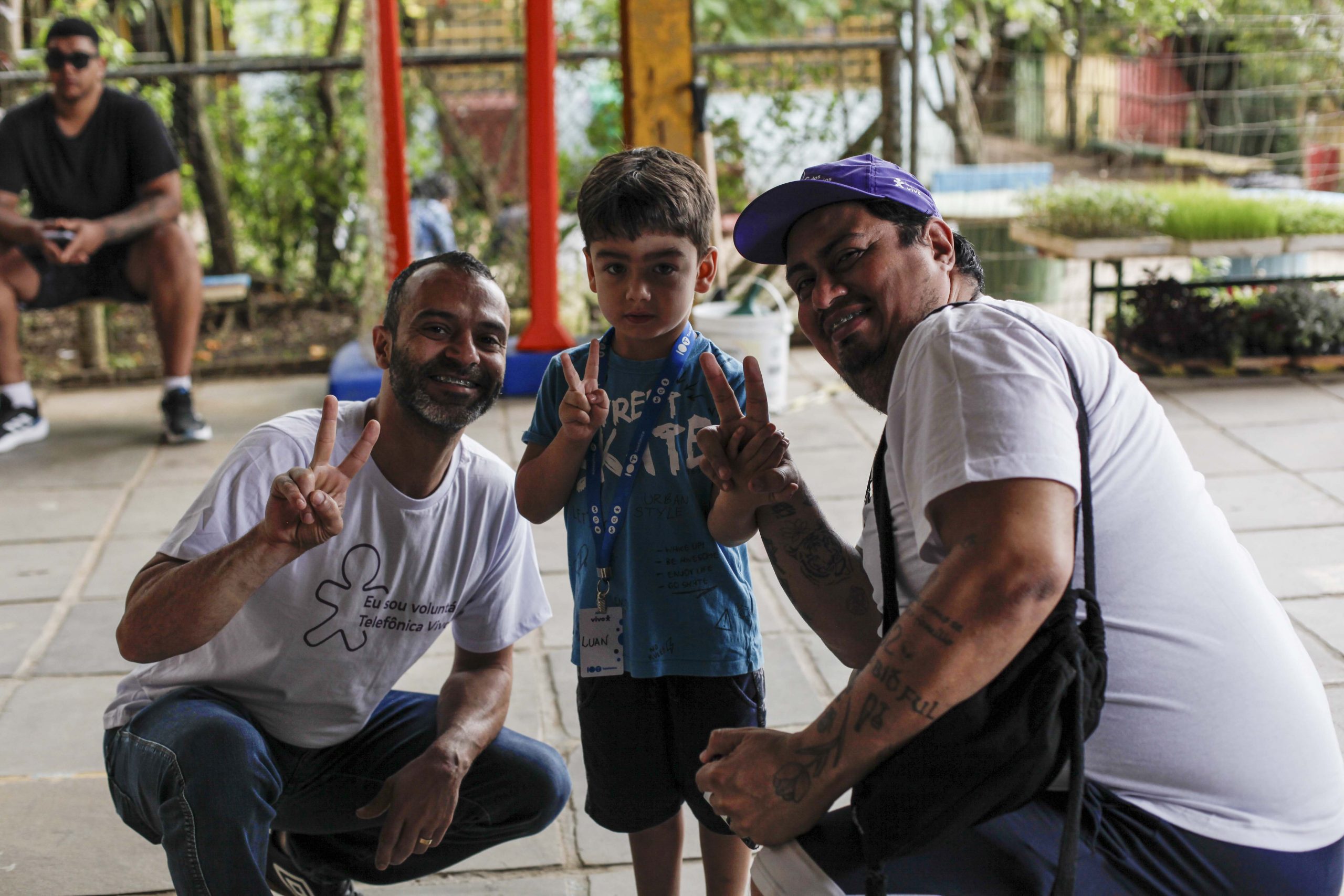 Dois voluntários posam agachados com uma criança em pé, todos fazendo sinal de vitória com as mãos.