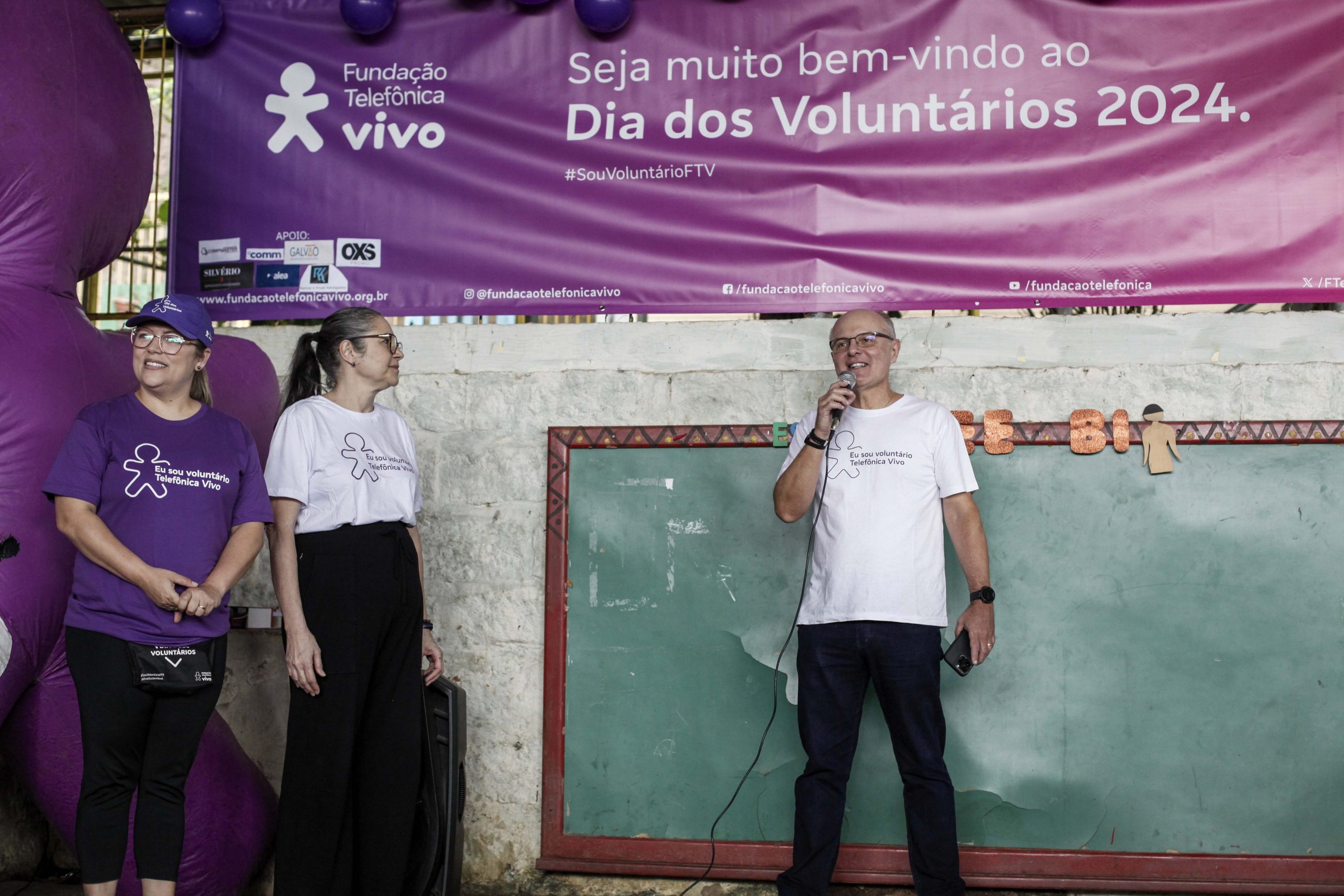 Voluntário fala ao microfone no palco, em frente a uma lousa, acompanhado de duas voluntárias.