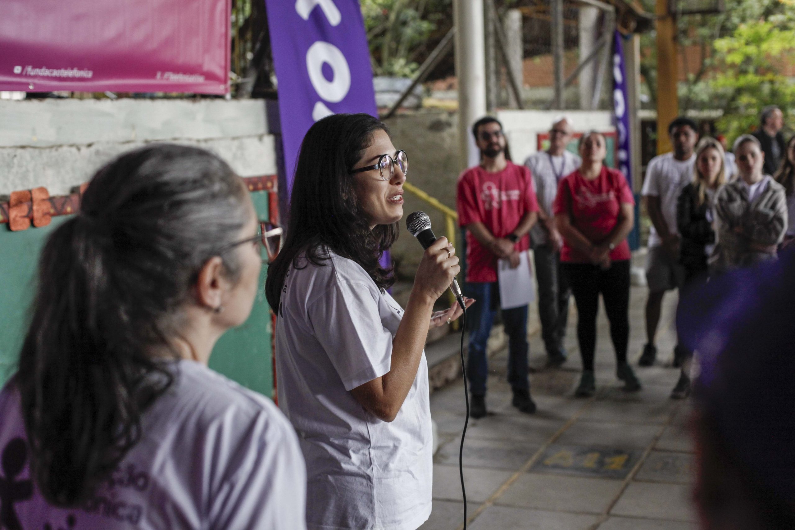 Voluntária fala ao microfone com multidão de voluntários no pátio da escola.